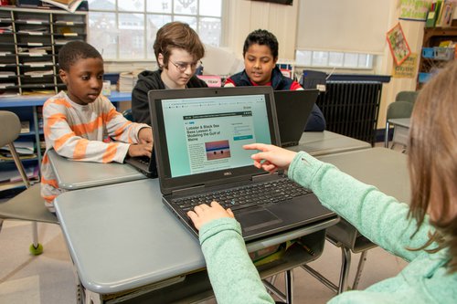 Students gather around two computers at classroom desks and learn about modeling.