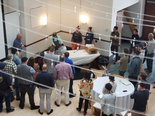 A lot of people are gathered in a large room, observing a man lecturing at a table with a tuna head on top of it.