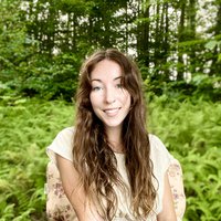 A portrait of a woman smiling in a white billowy shirt, surrounded by shrubbery.