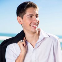 A portrait of a main holding his black jacket over his shoulder, standing on the sunny beach, and smiling in a white button down shirt.