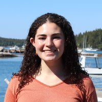 A portrait of a woman smiling for the camera. She has curly hair and a colorful shirt and is standing near a marina.