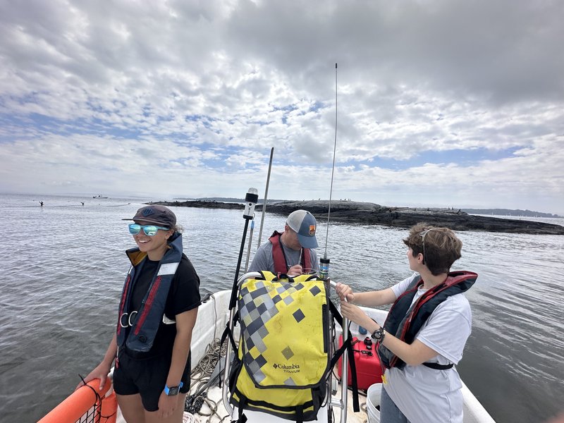 Two interns and a GMRI scientist stand on a skiff boat in the water.