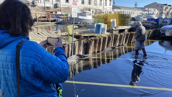 A woman takes a photo of a man walking in flooded waters on a parking lot.