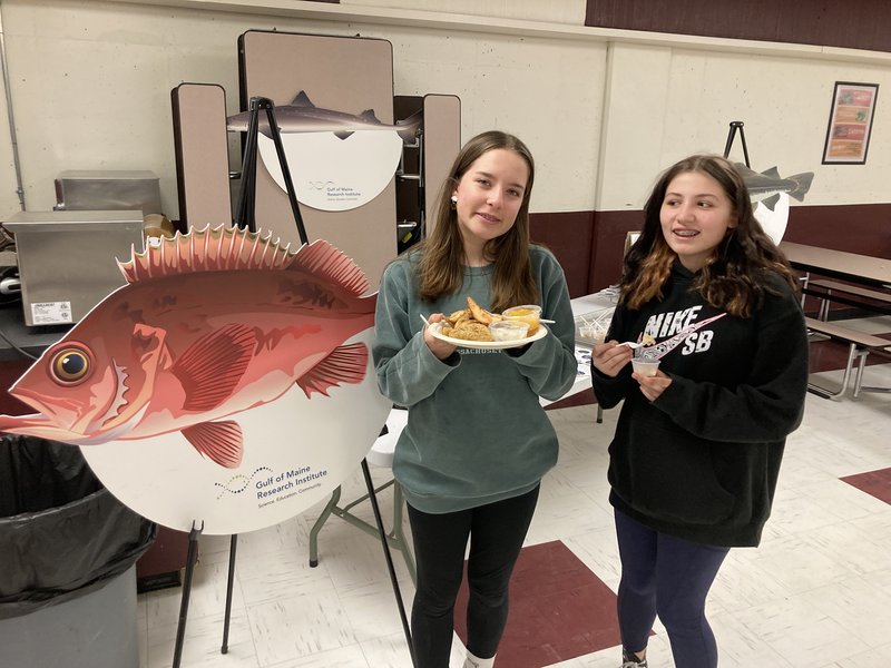 Two middle school students pose with dishes of seafood in front of a large poster of an acadian redfish.
