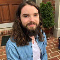 A potrait of a man with long black hair and a black beard sitting on a brick stoop with a denim shirt unbuttoned.