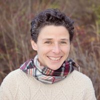 A portrait of a woman smiling with a plaid scarf and a beige sweater, and blurry brush behind her.