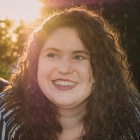 A portrait of a woman in a striped shirt with curly hair, smiling to the side of the camera as a soft yellow glow emerges behind her from the sun through trees.