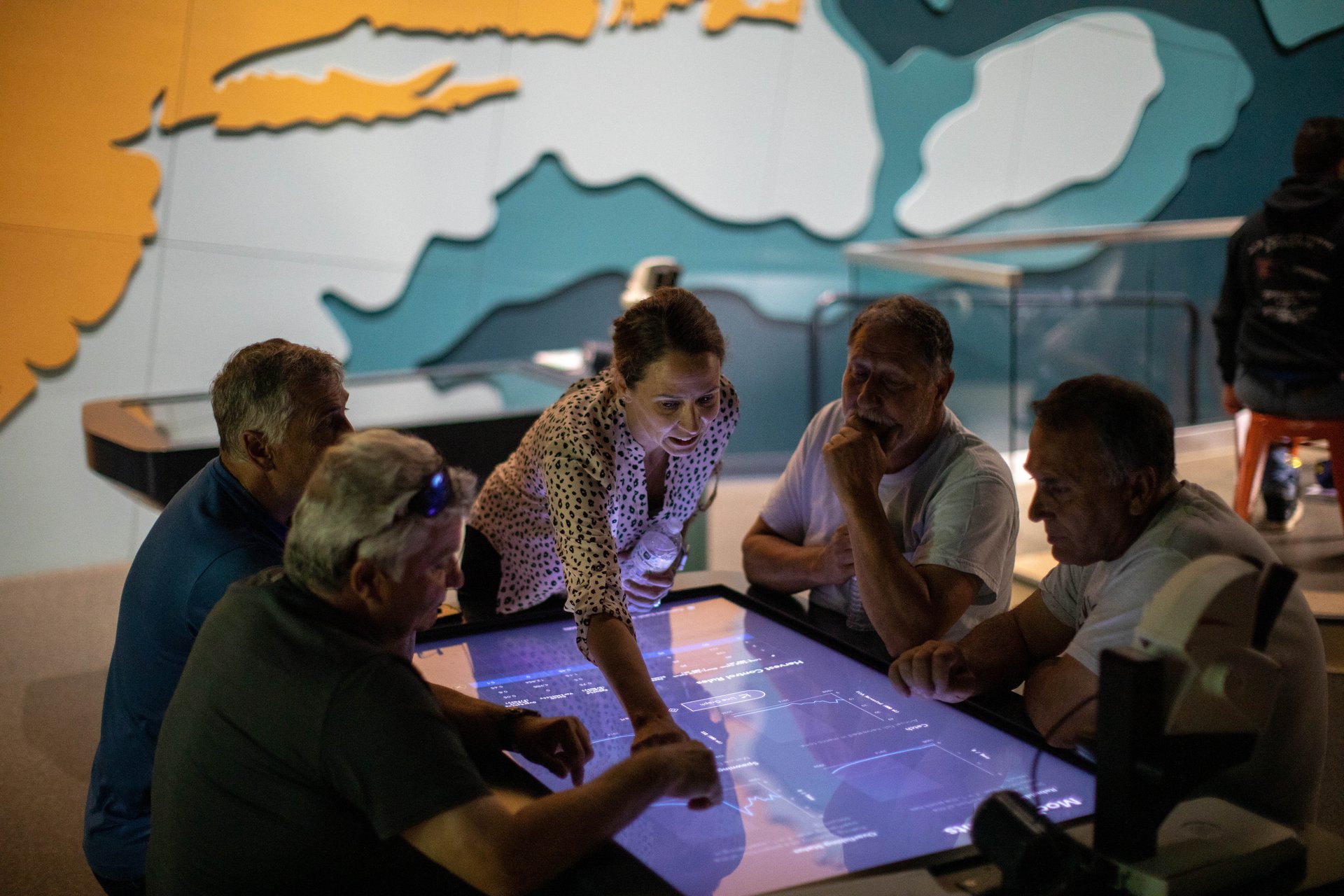 A woman engages a group of people around an interactive touch table in a dark-lit room.