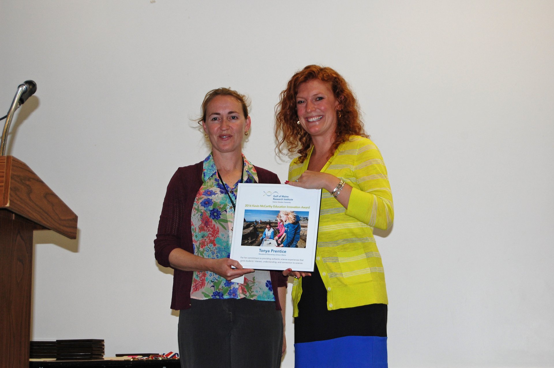 Woodstock Elementary School teacher Tonya Prentice holds up her McCarthy award in a flowery shirt, next to GMRI education staff member Meredyth Sullivan in a bright yellow shirt.