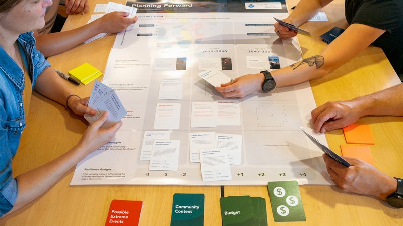 Four people sit around a table with cards in their hands, which are resting above something that looks similar to a board game.
