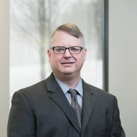 A portrait of a man in a dark grey suit, a light blue button down, and a grey patterned tie, with glasses on, smiling for the camera.
