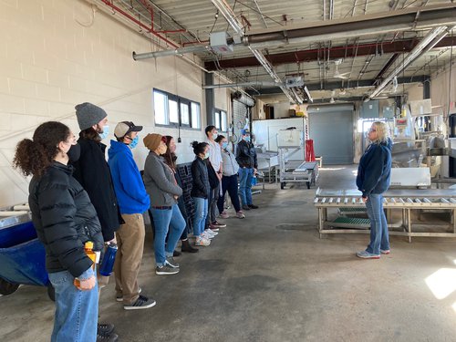 10 students stand in a line along the left side of the photo, facing a speaker talking to them who is standing on the right side of the photo. They are standing inside a Portland Fish Exchange building with concrete floors.