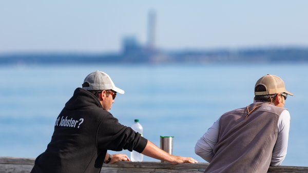 Two men with ball caps on are observing the ocean over a railing.