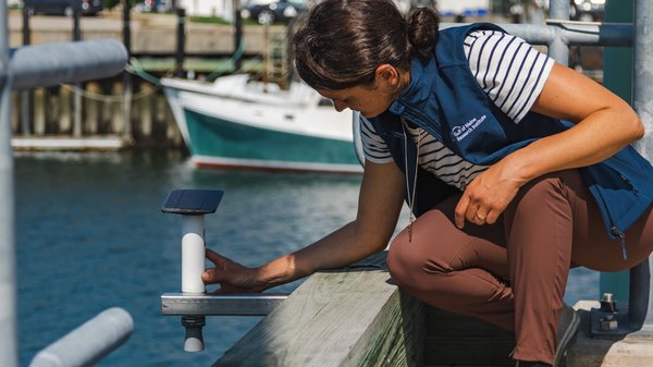 A woman in a blue vest and brown pants kneels down next to a rectangular, black device hanging out over a pier.