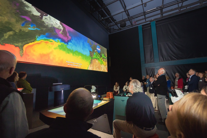 Adults gather in a large dimly lit room and look at a colorful digital display of the Gulf of Maine on a wall on the left side of the photo.