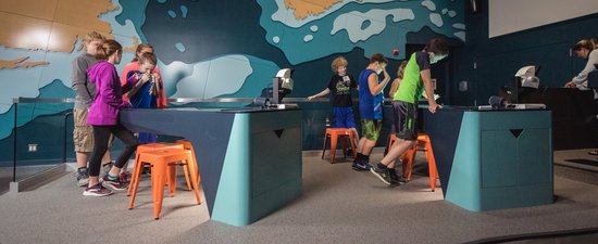 This image shows one area of the LabVenture facility, with students looking down at interactive, virtual kiosks in front of a large wall with a big map of the Gulf of Maine on it.