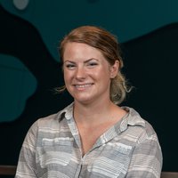 A woman smiles for the camera in a grey pullover while holding a black camera.