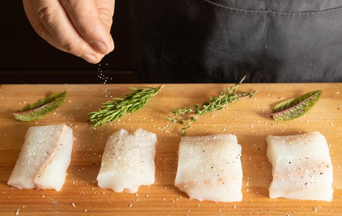 A chef sprinkles salt on fish fillets on a wooden cutting board with herbs next to the fillets