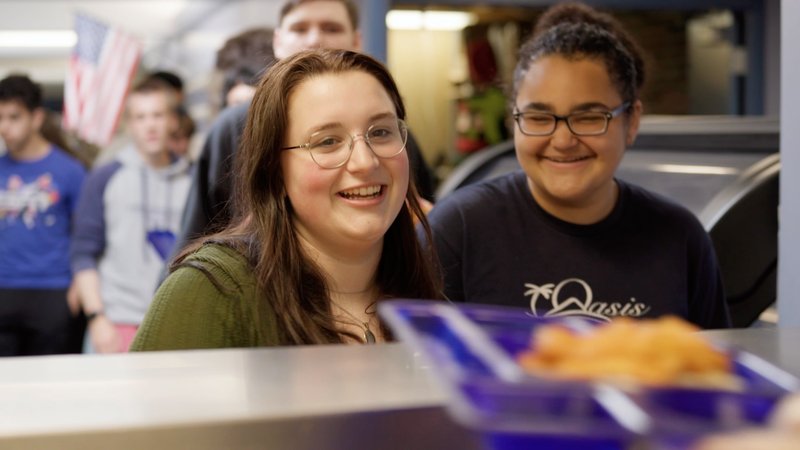 Two high school students waiting to be served seafood at school lunch.