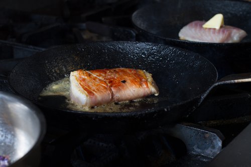 A rectangular piece of fish is browning on a black frying pan in the foreground, while a more raw piece of fish with a square of butter melts on top.