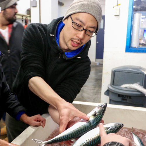 Japanese chef in a black zip up jacket and a gray hat holds a slender fish out to a customer in his right hand.