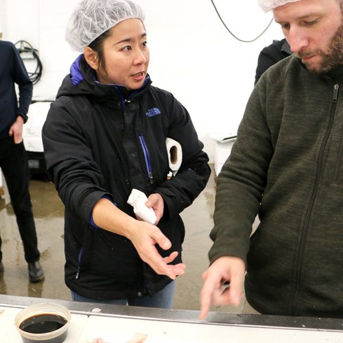 A Japanese woman in a black jacket and a white hair net holds her right hand out as she discusses seafood with a man in a green sweater and a white hairnet.