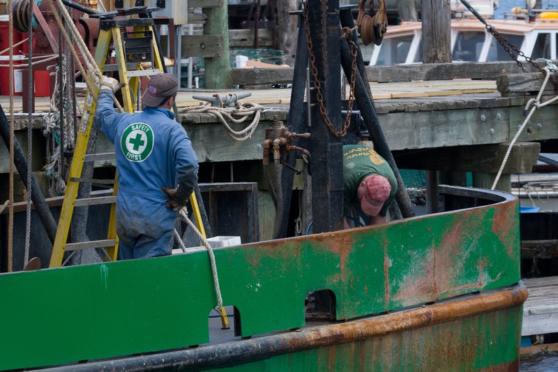 Two men attend to mechanisms on their green boat. Both are facing away from the camera, the man on the left is in blue and the circle on his back that reads 'Safety first" has a green background with white text.