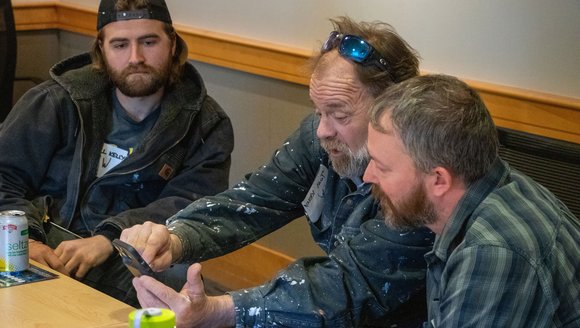 Three men are engaged in discussion at a table, looking at a phone.